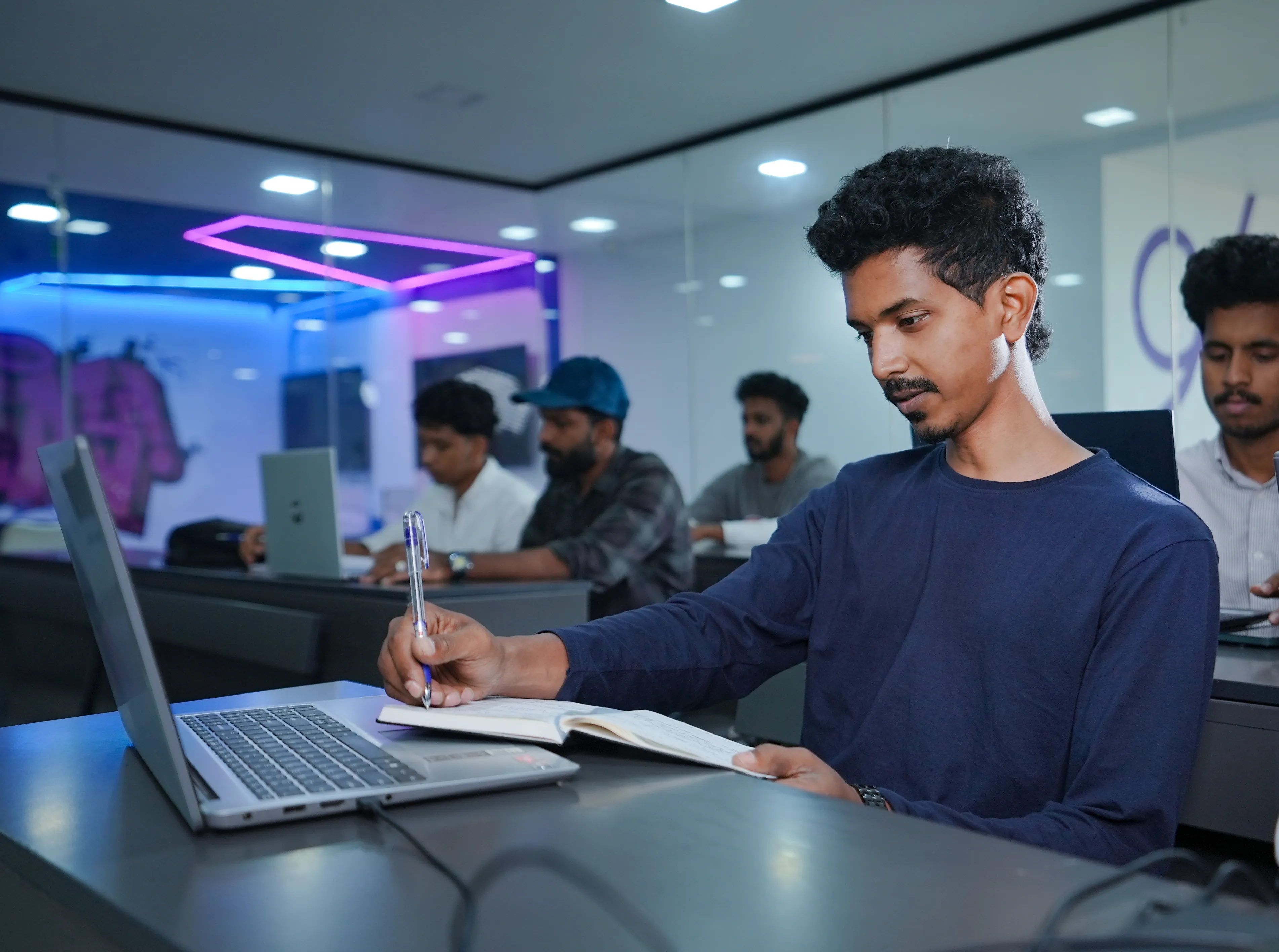 Student sitting at a desk, writing notes in a notebook while using a laptop, with other students working on laptops in a modern classroom setting with glass walls and colorful LED lighting.