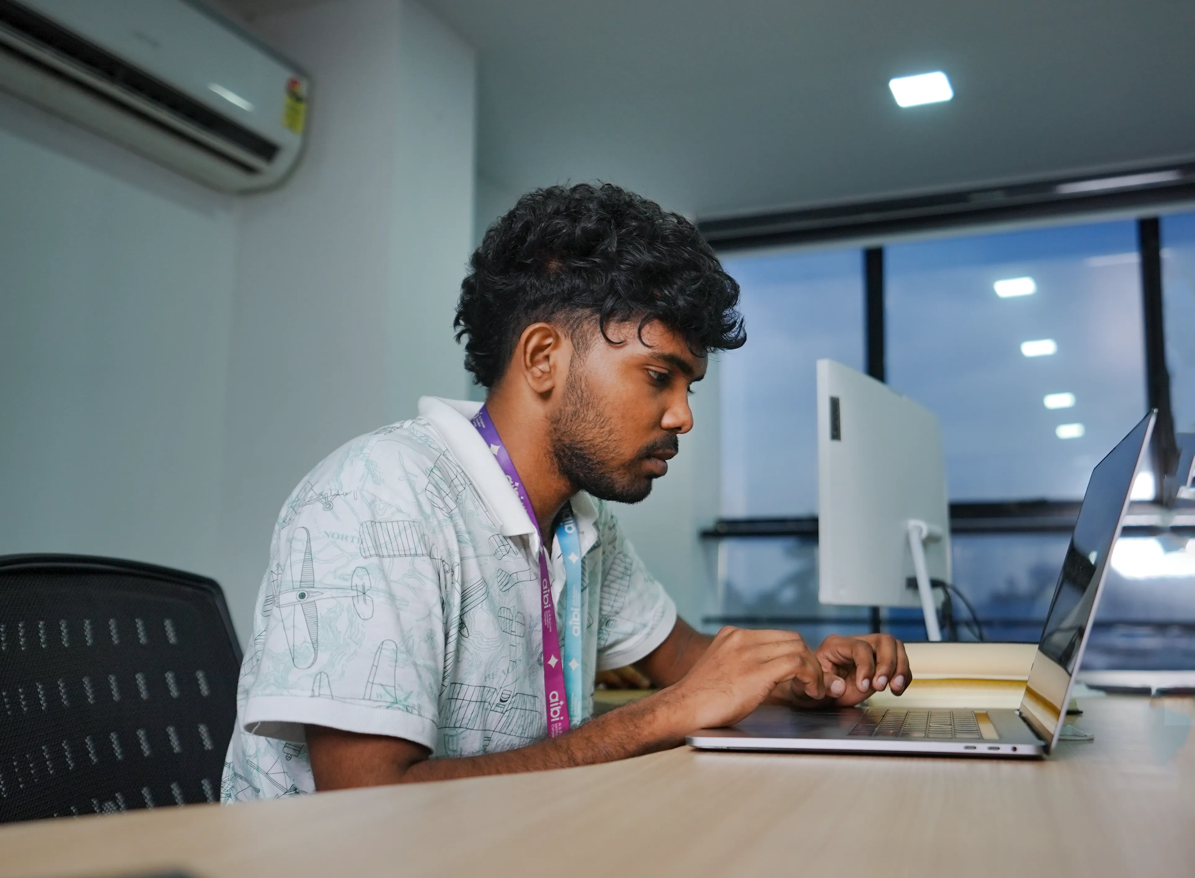 Student working on a laptop in a computer lab, focused on typing, with desktop monitors and a windowed office background visible behind him.