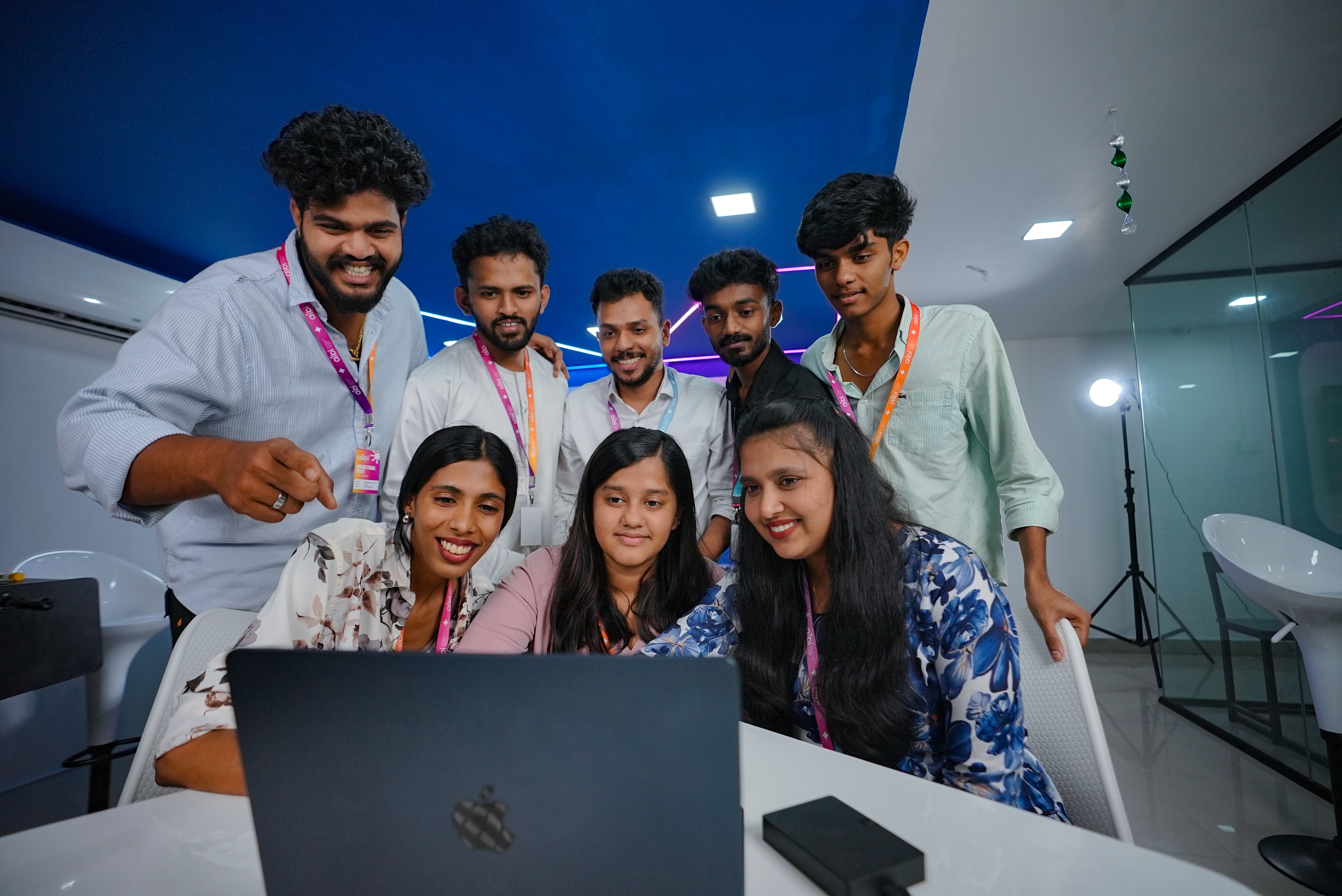 A group of young professionals wearing ID lanyards gather around a laptop in a modern office, smiling and discussing something on the screen. The room has blue ceiling panels, bright lighting, and a collaborative workspace setup.